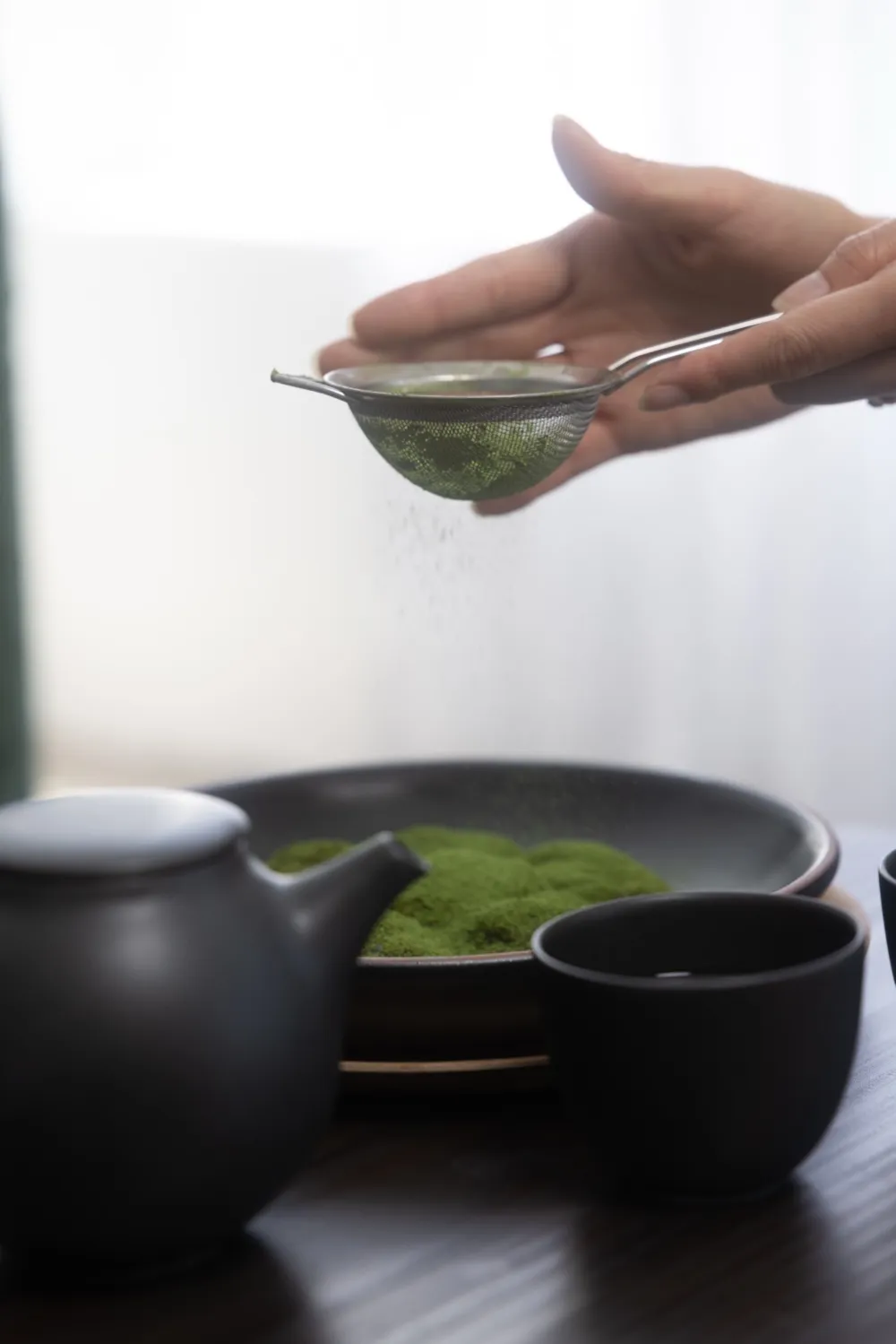Matcha being sifted over warabimochi beside a Japanese teapot